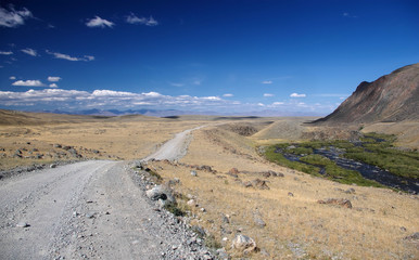 Road path on a desert wild mountain plateau with the yellow dry grass at the background of the hills under a blue sky with white clouds, Plateau Ukok, Altai, Siberia, Russia