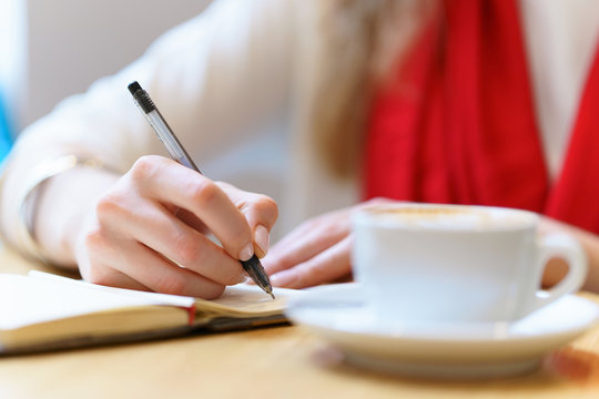 European woman with red scarf is writing by pen somthing in the notepad near white cup of coffee on table