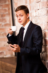 Young handsome businessman in black suit is drinking coffee and surfing in the phone near brick wall
