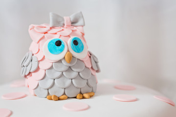 Detail of birthday cake covered with fondant displayed on the pink cloth and glass tray; decorated with pink dots and an grey and pink fondant owl with blue eyes sitting on top