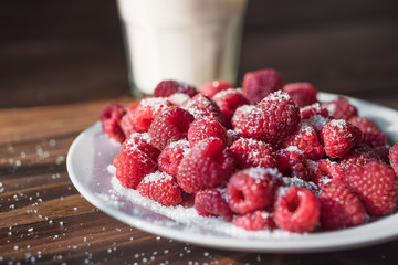 Ripe raspberry on a white plate powdered with sugar. Plate on a wooden table. Sunlight shines through the frame of a window. Glass of milk at the background.