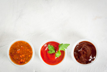 Three kinds of red tomato sauce on a white stone table: traditional classic ketchup, barbecue sauce, sweet and sour Chinese sauce. Top view copy space