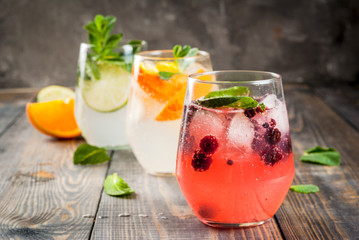 Selection of three kinds of gin tonic: with blackberries, with orange, with lime and mint leaves. In glasses on a rustic wooden background. Copy space 