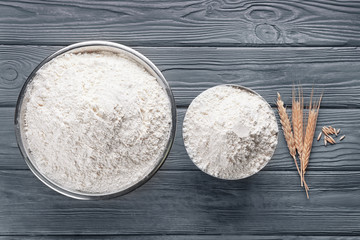 Two bowls of white flour and wheat ears on wooden background