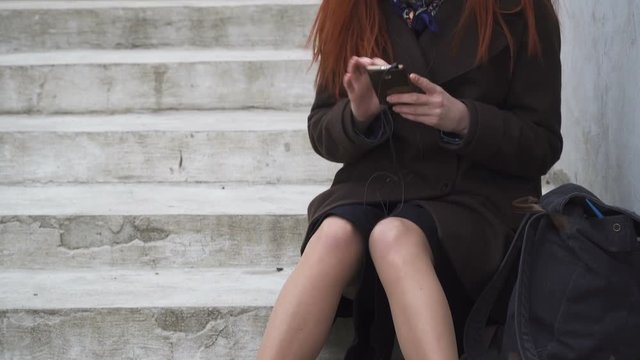 Cheerful woman is sitting on the stairs and listening to music, tapping the rhythm and flipping her smartphone, pan