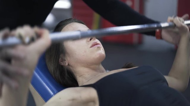 Close up of a young sweating woman lifting a barbell with a coach helping her. Locked down real time close up shot