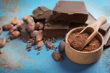Broken chocolate pieces, bowl with cocoa powder and beans on color background
