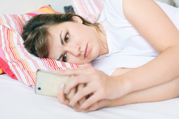 Young woman lying in bed and sending text message on the phone after waking up in the morning