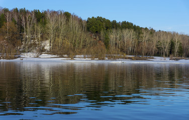 Obraz premium The river bank with trees and snow. Blue sky reflected in the water. Beautiful spring landscape.