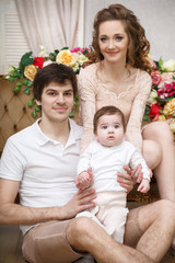 Mom, dad and baby are sitting on the couch in the background of flowers.