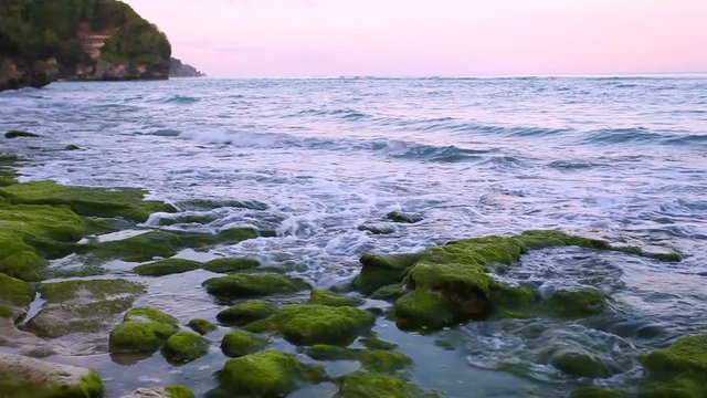 Calm ocean at sunrise, small waves wash over the rocks on the beach with mountains in the background. Bali Indonesia