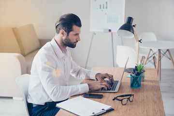 Side view of confident bearded concentrated businessman in white shirt works on laptop while...