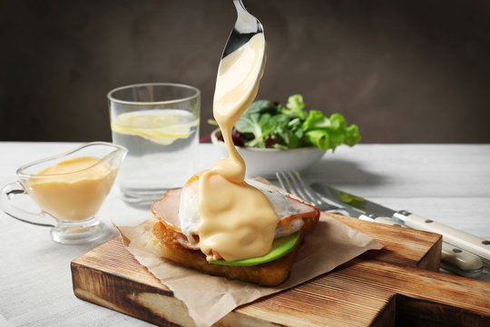 Pouring hollandaise sauce onto egg Benedict on wooden board