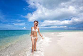 Portrait of a beautiful young woman in a white bikini walking along a beautiful tropical beach with azure water and white sand