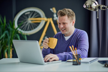 Young freelancer typing on keyboard and drinking coffee while sitting in home office. complementary colors used
