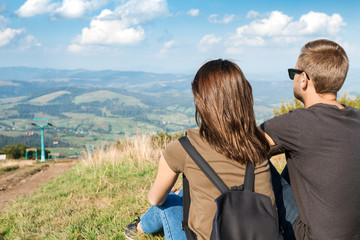 Young couple enjoying mountains landscape, sitting on hill back to camera.