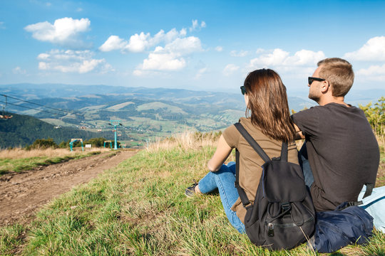 Young couple enjoying mountains landscape, sitting on hill back to camera.