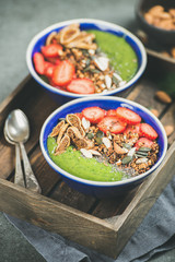 Healthy breakfast. Green smoothie bowls with strawberries, granola, chia and pumpkin seeds, dried fruit and nut in wooden tray over grey concrete background, selective focus. Clean eating food concept