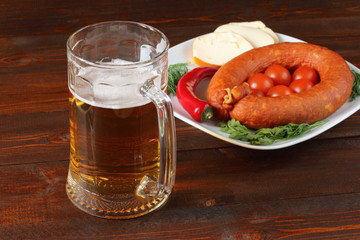 Mug of light beer and a plate with smoked sausage, cheese and vegetables are on dark wooden background.