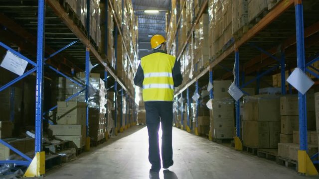 Following Shot of a Warehouse Worker Wearing Hard Hat Walking Through Rows of Storage Racks. Shot on RED EPIC-W 8K Helium Cinema Camera.