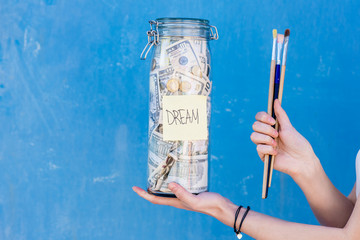 Woman holding a bottle full of money savings for the dream to be a painter on the blue background