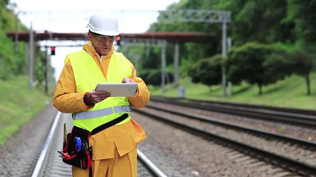 Railway employee in yellow uniform with tablet computer stands on railway line. Railway worker makes notes in his tablet computer. Railway worker in uniform and white hard hat with tablet pc in hands