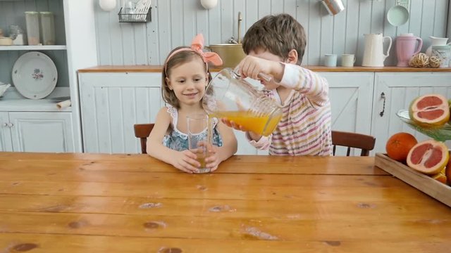 Boy brother serving orange juice for children girl sister in kitchen, happy family