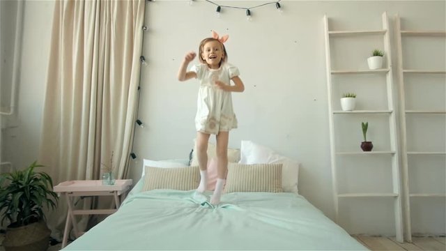 A happy cute young girl in white dress having fun jumping on bed, little smiling child