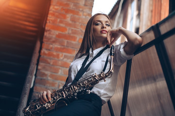 Young attractive girl standing in white shirt with a saxophone - outdoor. Sexy young woman with sax...