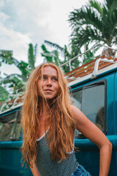 Red hair surf girl near blue vintage mini van with palm trees in the background in grey swimsuit, Indonesia, Bali