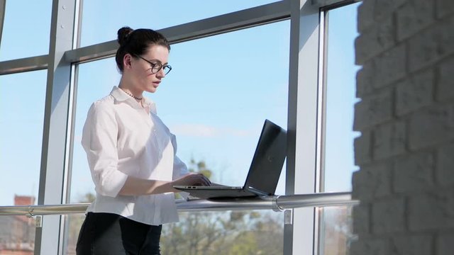 Young, pretty businesswoman working on laptop standing by window in office