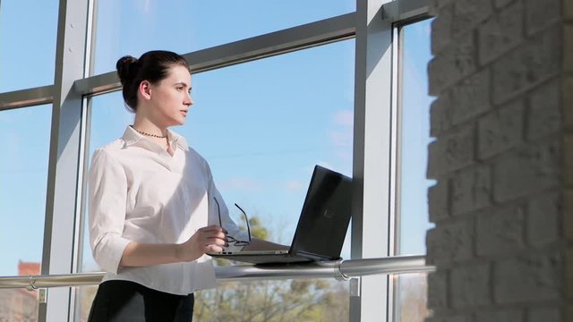 Young, pretty businesswoman working on laptop standing by window in office