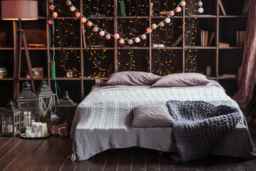 coziness, comfort, interior and holidays concept - cozy bedroom with bed and garland lights at...