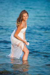 Beautiful woman standing in the sea in white dress. Pretty girl got wet her dress in the sea water. Attractive woman smiling at camera.