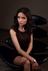 Sexy young model in brown dress sitting on the black chair on dark shadow background with fashion watch on the hand
