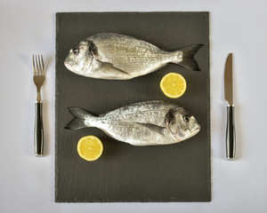 Two ready to cook raw bream fish with fork, knife and lemon on stone slate board. With copy space. Top view.