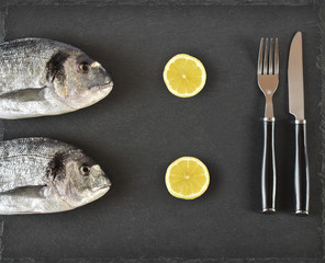 Two ready to cook raw bream fish with fork, knife and lemon on stone slate board. With copy space. Top view.