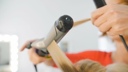 Hairdresser making curls on customer blond hair using electric curler in studio with white wall in background