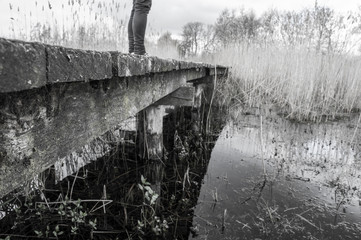 Black and white picture of a close up of the legs of a woman standing on an old wooden bridge over a little stream