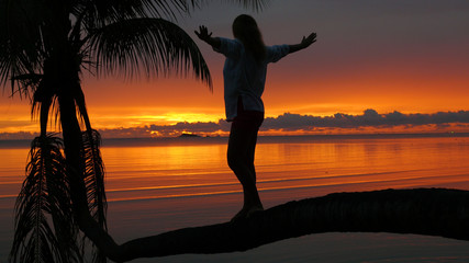 a girl stands on a palm tree on a background of red sunset on the seashore