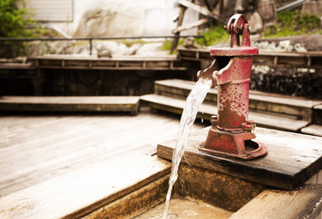 A small red vintage steel water pump mounted on a plank of wood with water pouring out of the spout