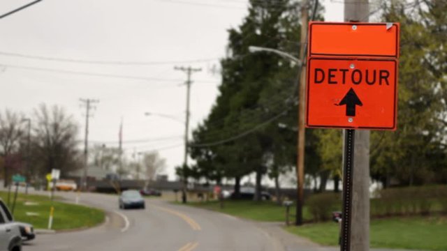 A orange construction detour sign with arrow pointing straight in generic overcasy residential road