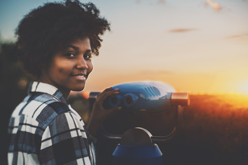 Cute biracial teenage female in plaid shirt is using street telescope to observe evening cityscape, young black girl with curly hair and dimples near blue binocular is preparing to look at sunset - Powered by Adobe