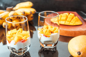 Glasses of yoghurt with mango and strawberry. Homemade healthy yogurt with fruits. A banch of bananas on a table