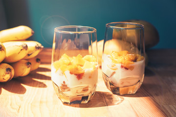 Glasses of yoghurt with mango and strawberry. Homemade healthy yogurt with fruits. A banch of bananas on a table. Morning sunlight