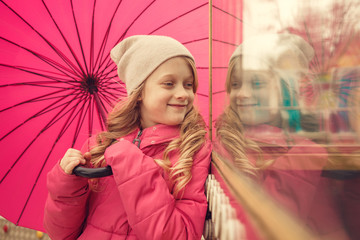 Cute little girl with umbrella looking on her reflection at window in park