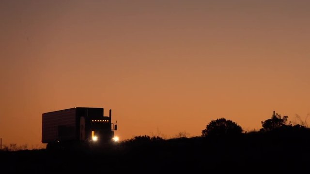 SLOW MOTION, CLOSE UP: Silhouetted freight container semi trailer trick traveling along the empty highway shipping cargo at peach-pink light of dawn. Transporter driving on the road hauling heavy load