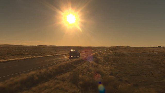 AERIAL: Flying above black SUV car driving on empty highway across beautiful remote wilderness in United States. People on road trip journey traveling across majestic Great Plains in golden light