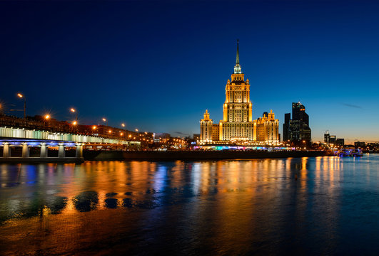 Amazing architecture view of Stalin tower illuminated in water and river Moscow shore with business skyscrapers at background. City landscape and dark blue sky after sunset, bridge and lights. Russia