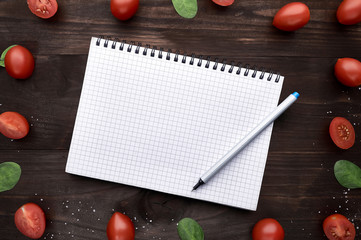 Notepad with a handle surrounded by sheets of arugula and small tomatoes with salt on a wooden background. Top view